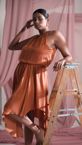 vertical video: model in elegant orange dress posing confidently on wooden ladder in studio