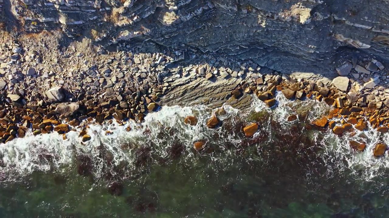 Ocean waves crash against rocky shore under clear sky along coastline during midday sunlight with patterns in the sand and stones visible
