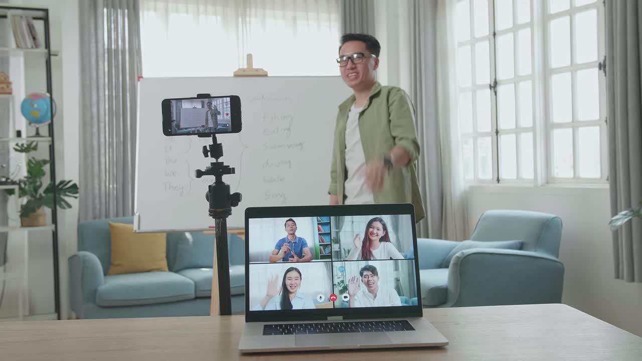 Asian Male Teacher With Glasses Shoots Video By Smartphone Camera And Having Video Call On Laptop While Teaching English At Home