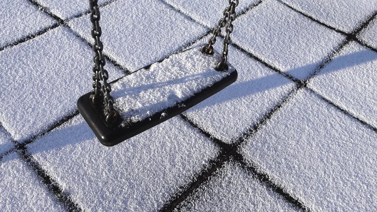 Top down slow motion video of a empty playground swing swinging and moving back and fourth during first snow day with white fluffy hale grainy snow around and on it. The swing has linked chain. Sunny