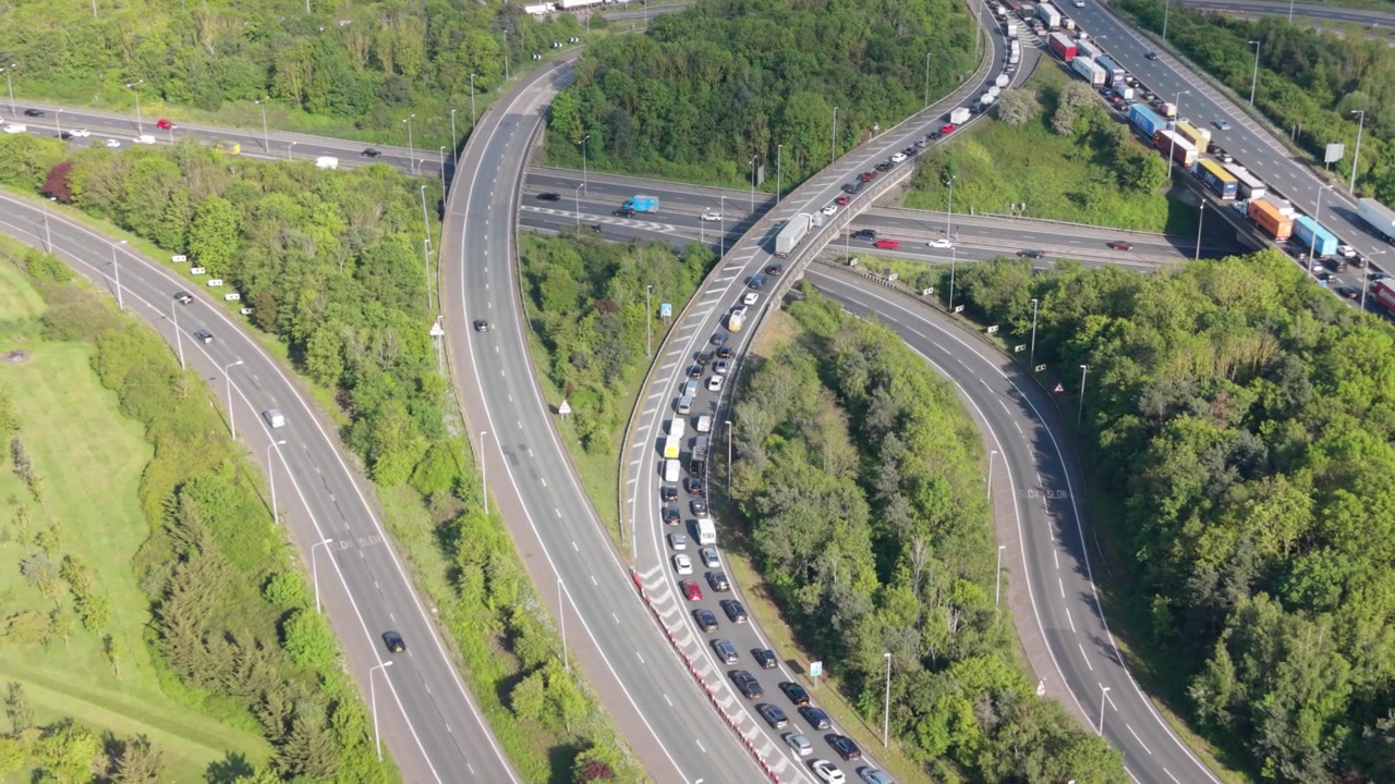 Yoğun bir otoyol kavşağının gündüz havadan görünümü. Bir yolda çok sayıda araba ve kamyonun biriktiği uzun bir trafik sıkışıklığı oluşmuş. Diğer bağlantı yollarında hareketli araçlar görülüyor.