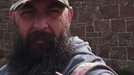 troubled bearded male wearing baseball cap standing next to stone wall looking away