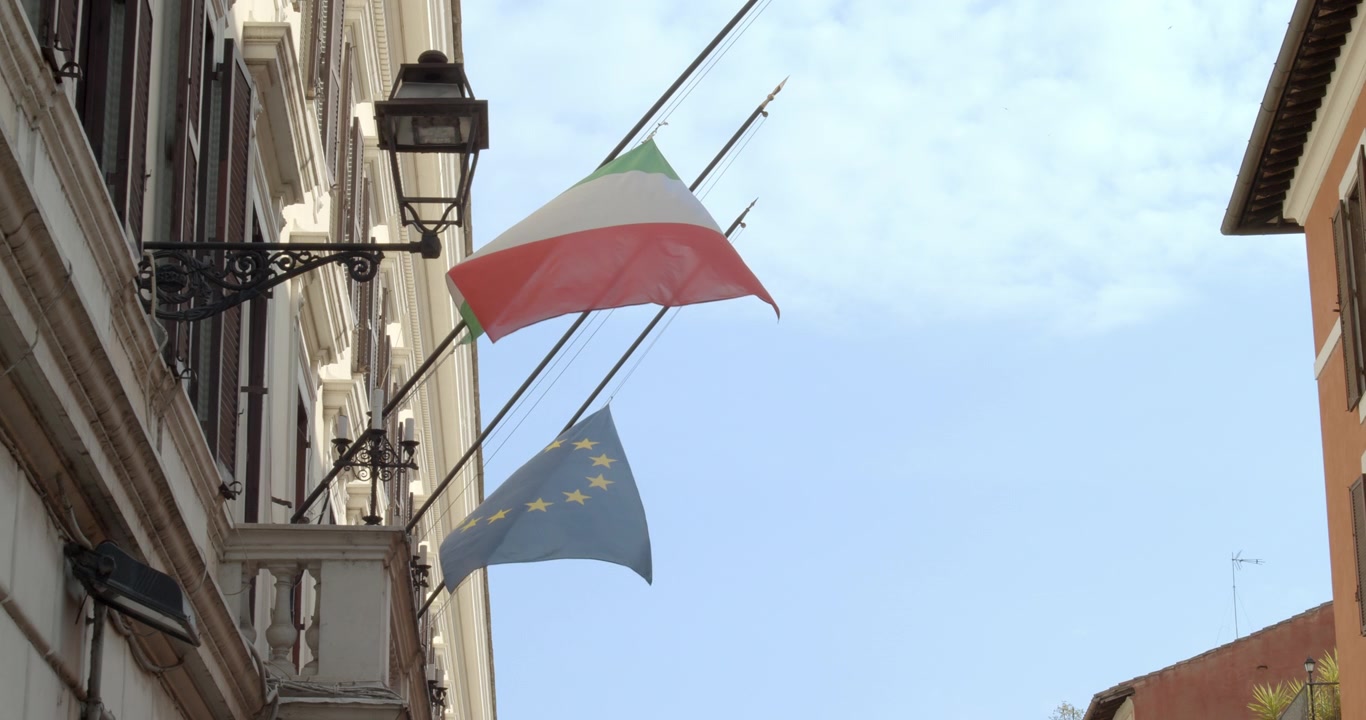 Bandiera italiana e bandiera europea sventolano al vento al rallentatore su un edificio italiano.