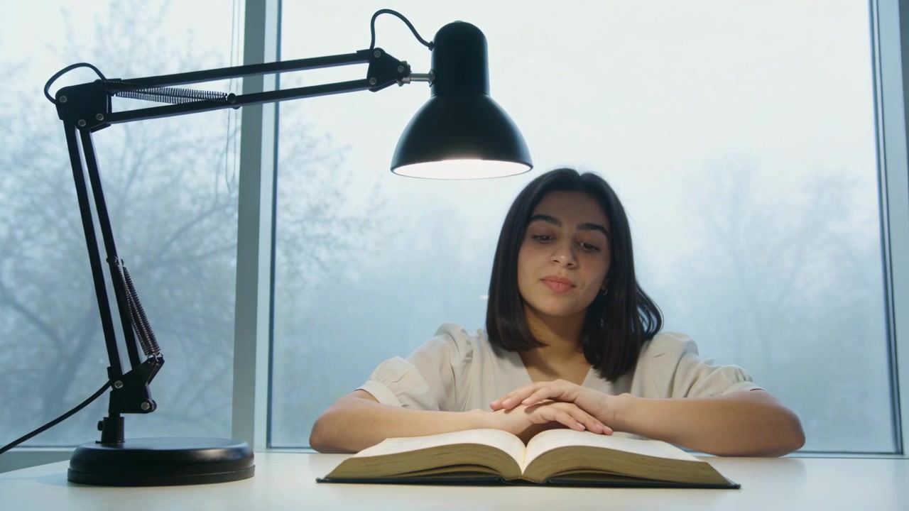 Reaching woman in light blouse adjusting lamp at desk, illuminating open book for reading, pausing