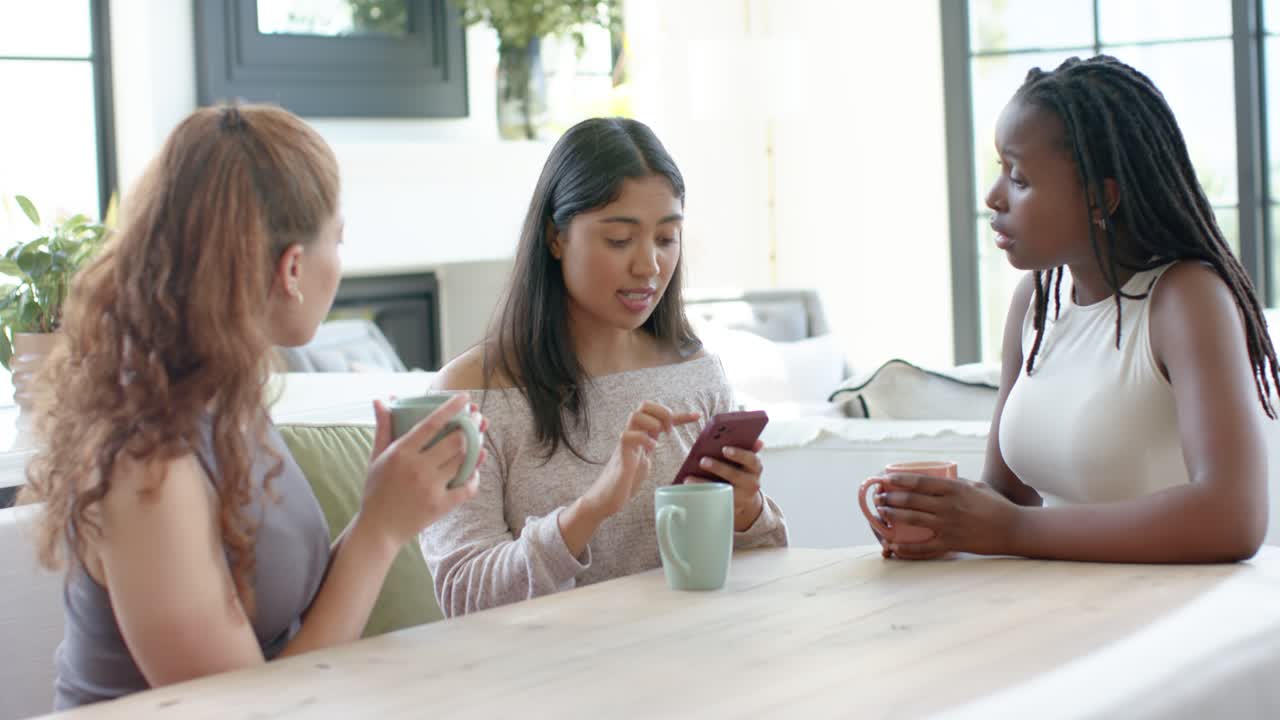 Diverse female friends at table watching center woman tapping smartphone, showing content with mugs