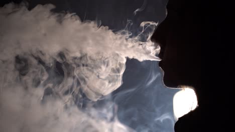 slow motion isolated silhouetted closeup profile view of a man smoking and blowing out a lot of smoke in dark background