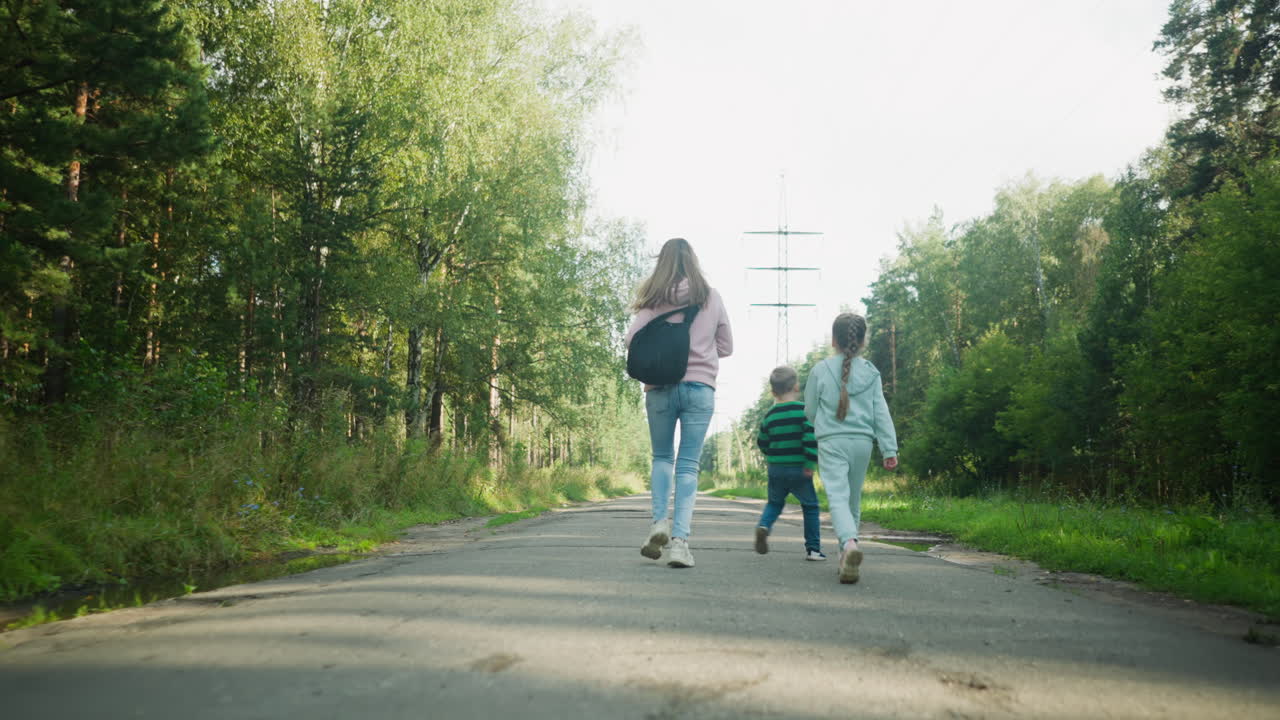 Rear view of joyful family running playfully down tarred road through serene forest area, with kids and older sister enjoying outdoor moment, surrounded by tall trees and soft morning sunlight