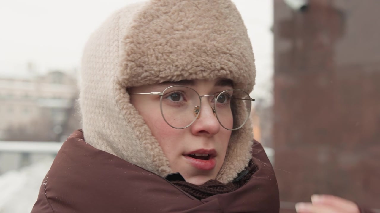 Three-quarter close-up of young person wearing fur-lined winter hat, glasses, and padded coat adjusting collar with hand while turning head sideways; pursed lips, flushed cheeks, and visible breath