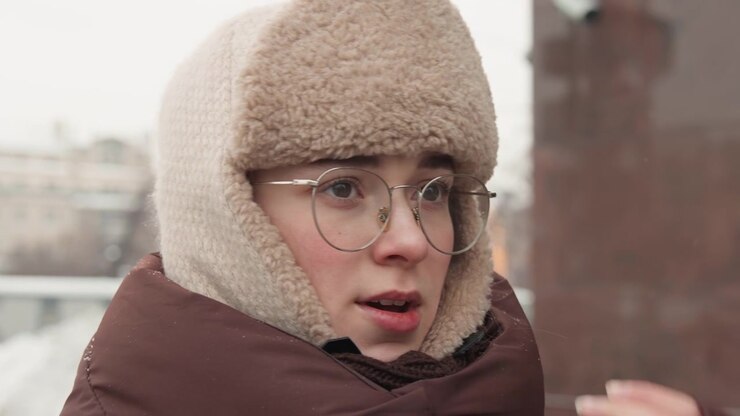 Three-quarter close-up of young person wearing fur-lined winter hat, glasses, and padded coat adjusting collar with hand while turning head sideways; pursed lips, flushed cheeks, and visible breath