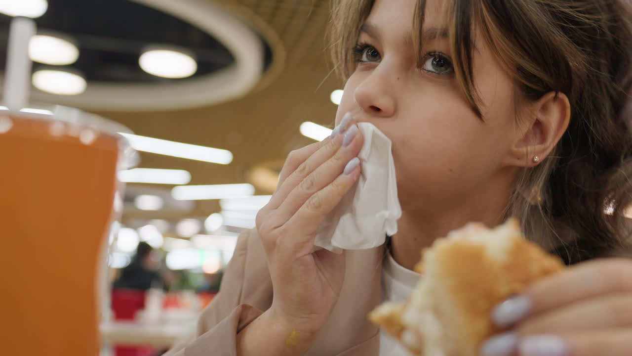 Jóvenes comiendo hamburguesas, persona joven masticando un sándwich crujiente en la cafetería de un centro comercial, adolescente mordiendo casualmente una hamburguesa crujiente en medio del bullicioso ambiente del patio de comidas del centro comercial