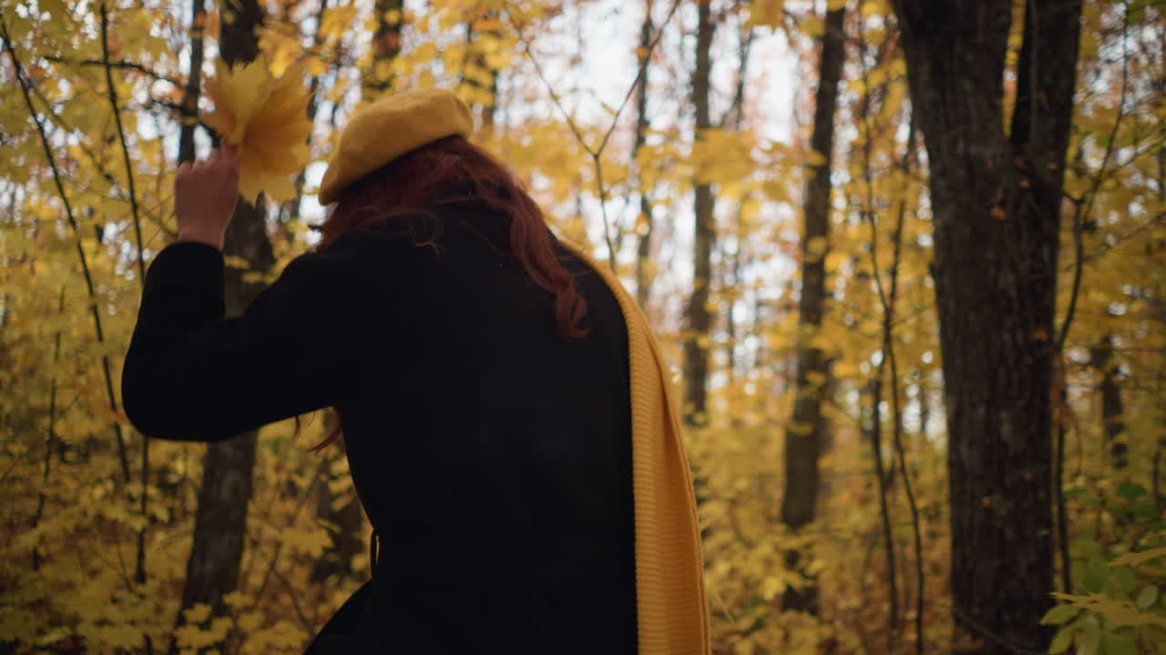 vista trasera de una mujer con boina amarilla y abrigo negro caminando por el bosque de otoño, con hojas doradas en la mano, rodeada de follaje vibrante, explora la naturaleza con serena curiosidad