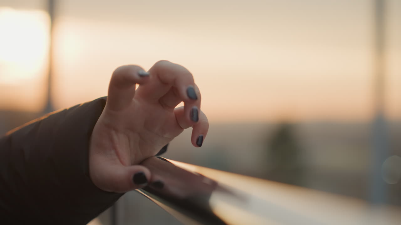 Bagliore del tramonto con unghie dipinte. Morbido bokeh cattura un tocco riflessivo durante la danza dell'ora d'oro. Primo piano di mano appoggiata su ringhiera al tramonto con unghie nere dipinte e delicata illuminazione bokeh.