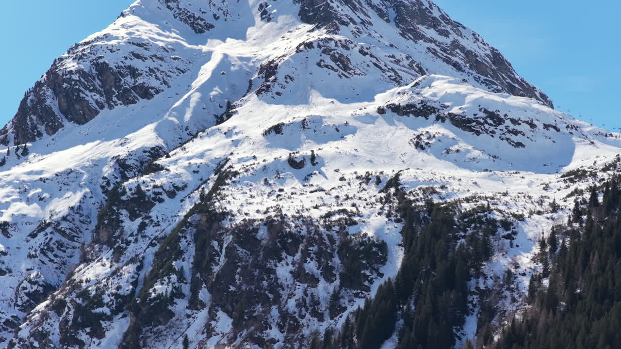 Panoramica aerea verso l'alto in primo piano di un'imponente montagna innevata in una giornata di sole.