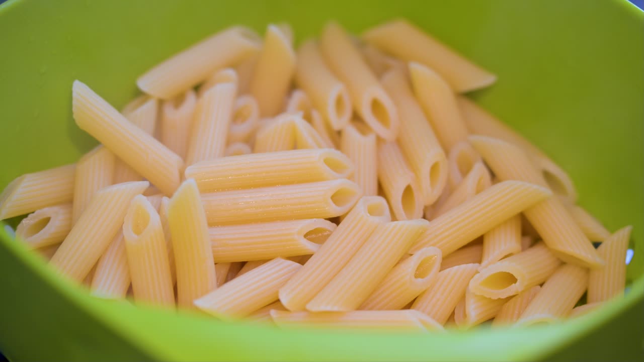 This close-up shot shows hot penne pasta in a green kitchen strainer. Steam is visible rising from the pasta after it has been boiled, ready to be served