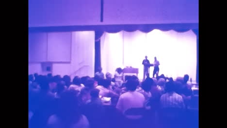 man standing on stage, addressing crowd at 1970s usa car show. circa usa - 1970s: a man confidently stands atop a stage, speaking to a lively crowd at an old car show in the 1970s usa.
