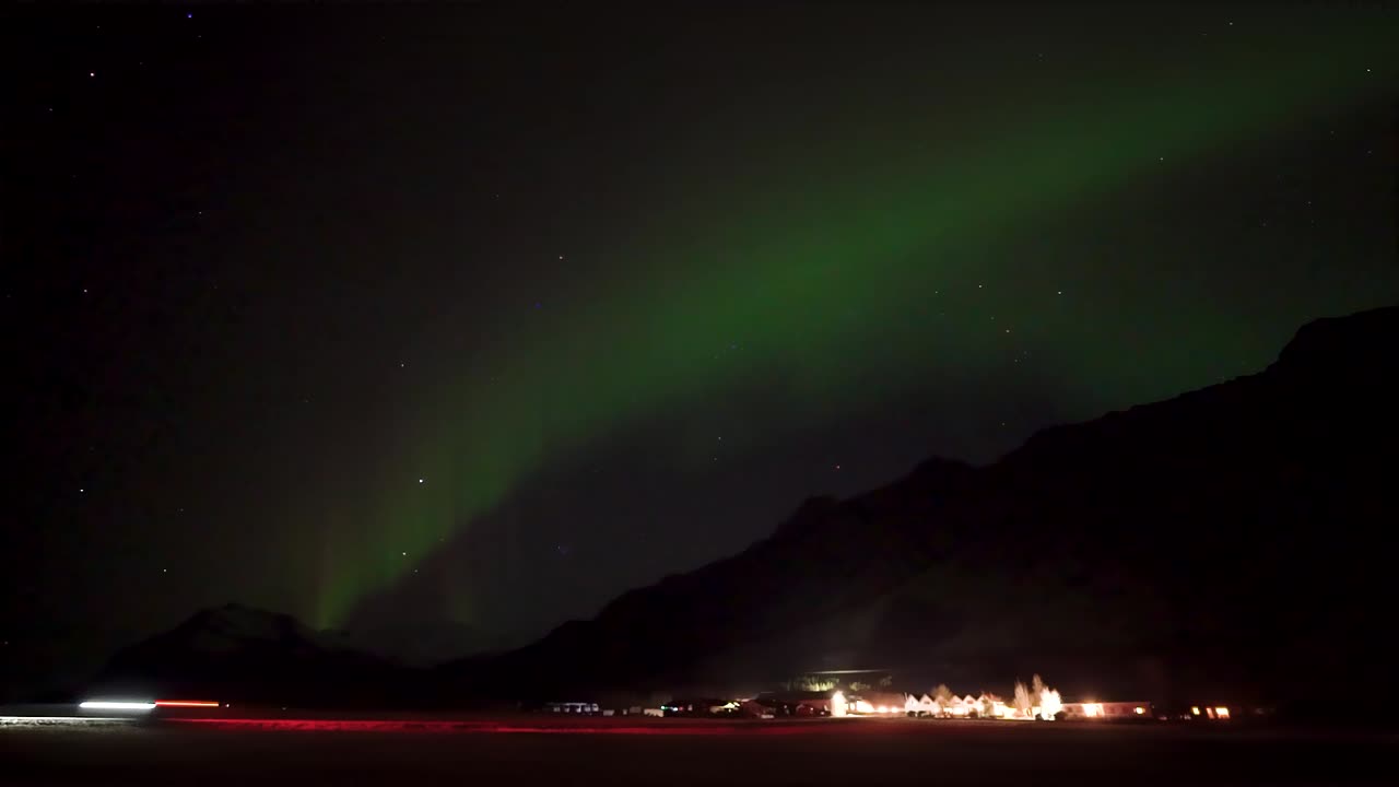 Green Aurora Borealis Illuminating Dark Night Sky Above Shadowy Mountains With Lit Buildings In Hali, Iceland. timelapse