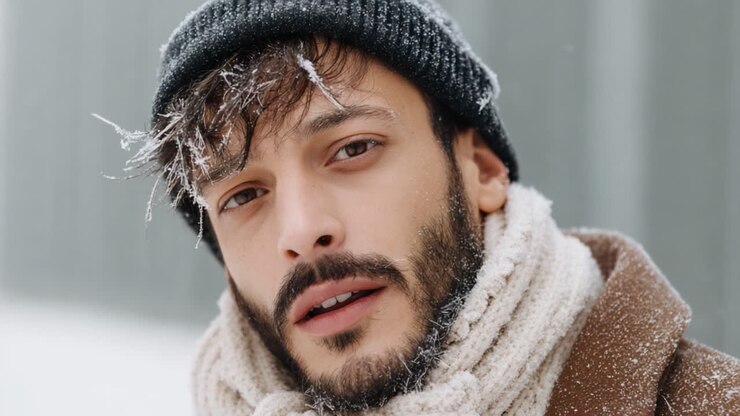 Primo piano di un giovane uomo circondato dalla neve invernale, che mette in mostra il suo elegante cappello e la sua calda sciarpa in un ambiente freddo e tempestoso