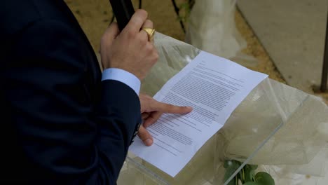 un hombre lee un texto en un plexiglas transparente en una boda, es un discurso, centrarse en la hoja y el dedo corriendo sobre las líneas, la mano que sostiene el micrófono