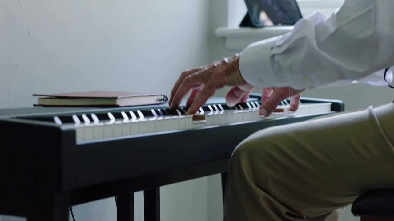 Le mani iniziano a muoversi, un pianista si esercita a casa in camicia bianca, premendo i tasti e il pedale del sustain.