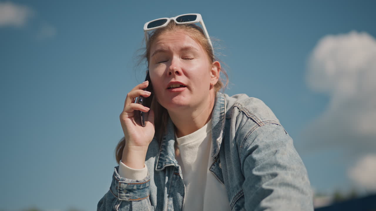 Donna bianca sollevata che riceve buone notizie, giacca di jeans con occhiali da sole appoggiati sulla testa, sorridente mentre tiene il telefono all'orecchio, reazione gioiosa ed espressiva, cielo soleggiato con nuvole soffici, celebrazione all'aperto.
