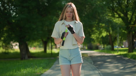 young woman strolls through a sunlit park, inspecting green air pump with focused expression, background includes trees and greenery, while another person passes by in distance