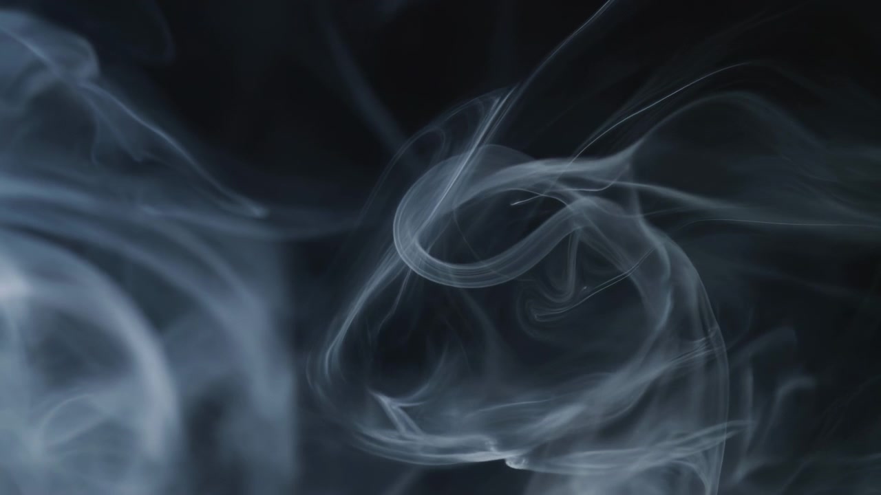 Movimento ravvicinato di fumo grigio-blu pallido che si trasforma da un anello strettamente arricciato in nastri aperti e fluidi e volute separate su uno sfondo scuro e profondo con un morbido contrasto luminoso, includendo fumo e nastri.