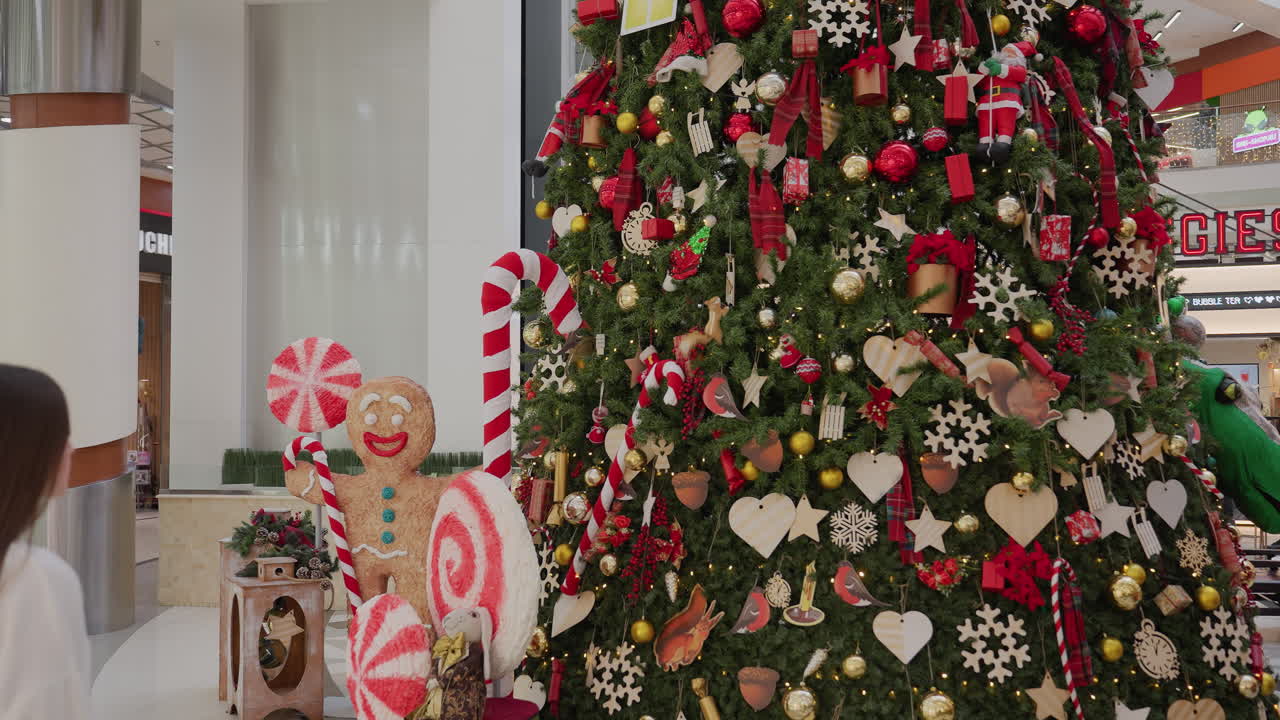 Back view of woman walking towards festive holiday tree with, Christmas decorations inside modern shopping mall, beautiful ornaments, candy canes, and gingerbread man near Christmas tree