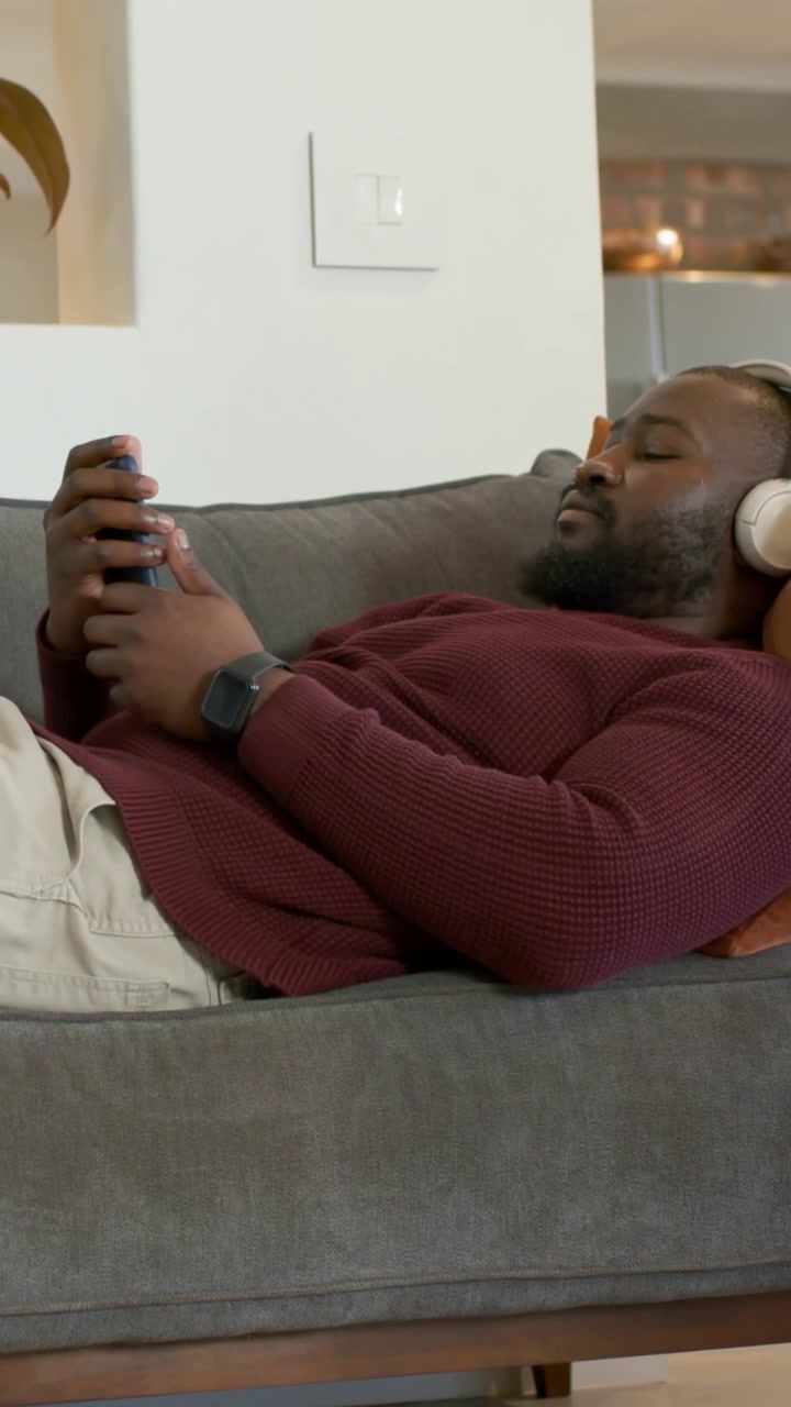 Video verticale: Uomo in maglione bordeaux che tocca un dispositivo, sistema le cuffie e si accomoda sul divano a casa per riposare.