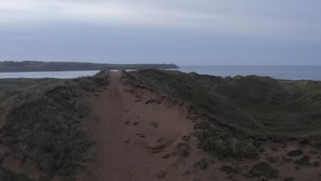 Twilight-aerial-flies-over-grassy-sand-dunes-toward-calm-ocean-beach