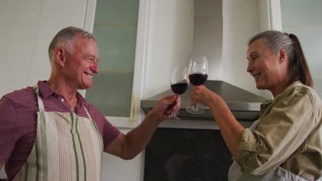 pareja de ancianos caucásicos usando delantales brindando y bebiendo vino en la cocina en casa