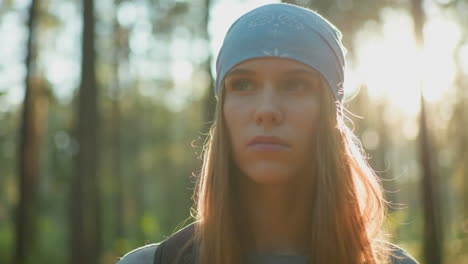 young woman in blue headscarf stands in forest, looking intense and contemplative as sunlight creates a radiant glow around her face and effect around from the background