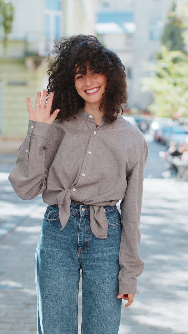 mujer joven sonriendo amigablemente a la cámara, agitando las manos hola, hola, saludo o adiós en la calle de la ciudad