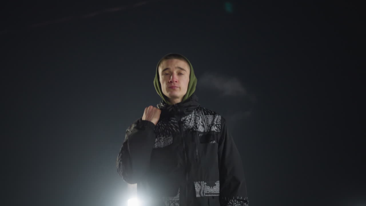 boy in green hood and patterned jacket walks through ice rink holding skate boot over shoulder with bright arena lights shining in background