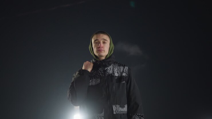 boy in green hood and patterned jacket walks through ice rink holding skate boot over shoulder with bright arena lights shining in background