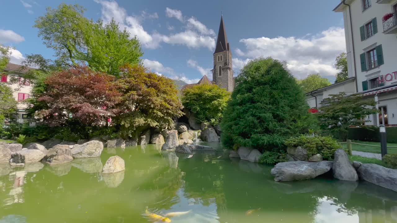 スイスのインターラーケンにある、緑の池を泳ぐ錦鯉、岩、豊かな木々、そして一部曇り空の下にそびえる教会の尖塔が特徴の、静かで穏やかな日本庭園の固定ショット。