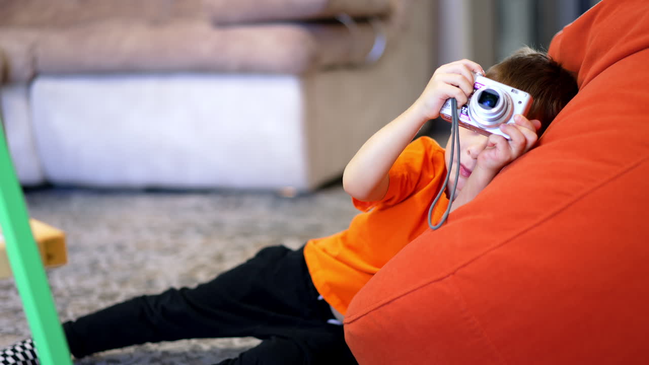 Lovely toddler taking pictures with an old-fashioned camera. Kid wearing orange t-shirt plays with photo camera at home.