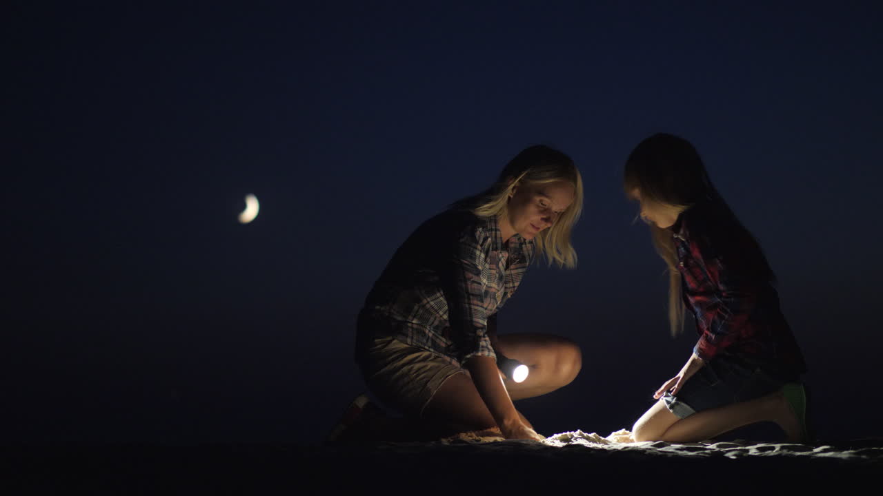 mamá e hija están jugando juntas en la arena por la noche brillan con una linterna buscando