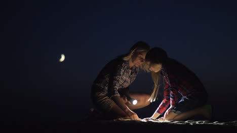 Mama-Und-Tochter-Spielen-Nachts-Zusammen-Im-Sand,-Sie-Leuchten-Mit-Einer-Taschenlampe-Auf-Der-Suche-Nach