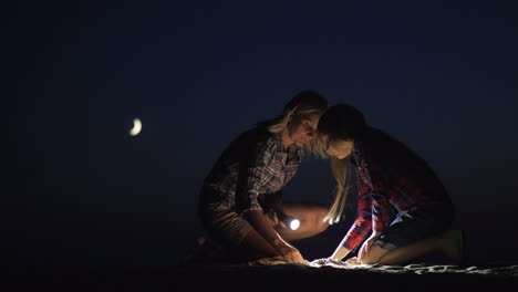 mom and daughter are playing together in the sand at night they shine with a flashlight looking for