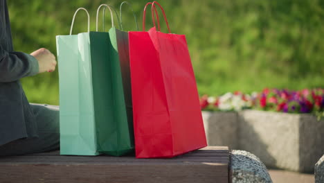 close-up of an individual dropping colorful shopping bags on a bench outdoors, the scene features a blurred background with vibrant flowers visible, and the person sits down in a relaxed manner