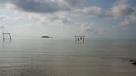 Schwarze-Silhouette-Von-Zwei-Kindern,-Die-Auf-Einer-Holzschaukel-Mitten-Im-Meerwasser-Bei-Karimun-Jawa-Spielen-Und-Stehen