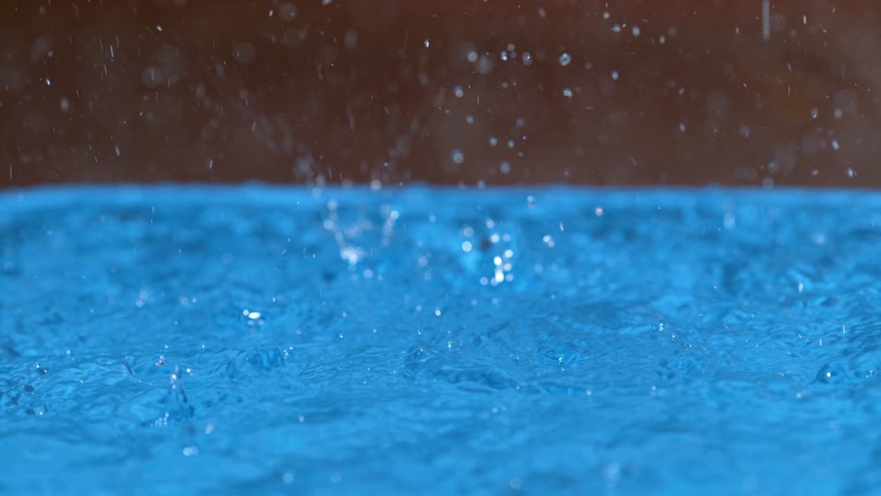 MACRO, DOF: Glassy droplets of rain fall from the sky and into the blue pool.