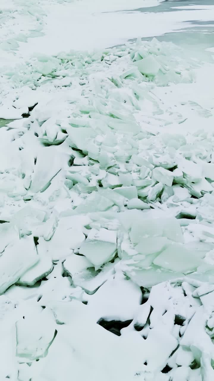 Primo piano di una cresta di pressione coperta di neve con lastre turchesi e ghiaccio marino frantumato accanto a una pozza di acqua di disgelo pallida, creando una robusta texture invernale minimalista in un'inquadratura verticale.