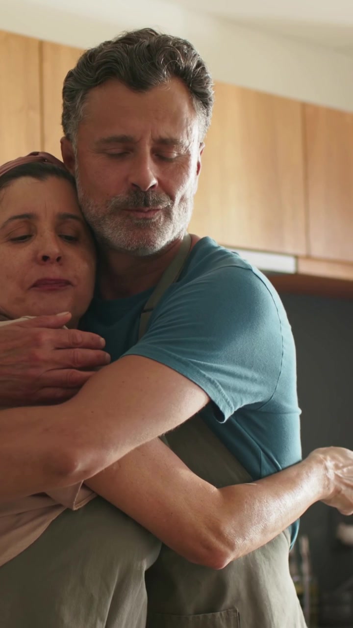 Video verticale: Uomo che abbraccia e conforta una donna da dietro, in una cucina domestica, entrambi con grembiuli.