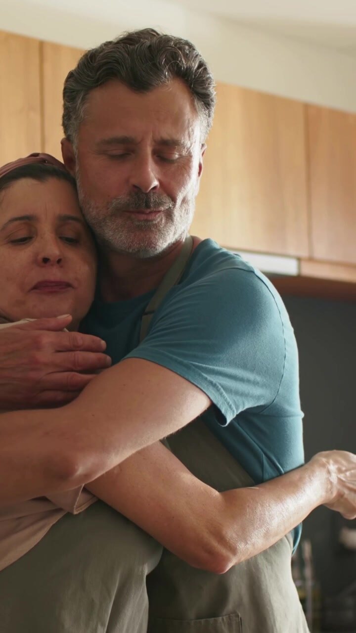 Video verticale: Uomo che abbraccia e conforta una donna da dietro, in una cucina domestica, entrambi con grembiuli.