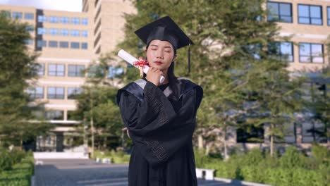 asian woman student graduates in cap and gown with diploma thinking about something and looking around in front of a magnificent university building