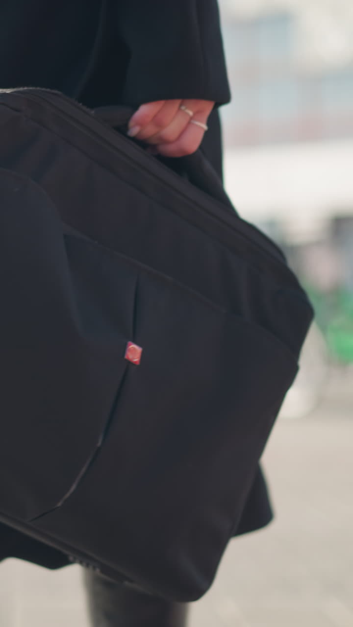 Close-up of black bag carried by person wearing stylish black coat and leather boots, walking confidently on sunny urban street with blurred modern architecture and green bikes in the background