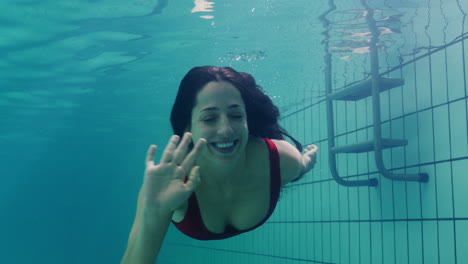 Hermosa-Mujer-Nadando-Bajo-El-Agua-En-La-Piscina-Sonriendo-Saludando-Con-La-Mano-Mujer-Atractiva-Disfrutando-Nadando-En-Agua-Clara-4k