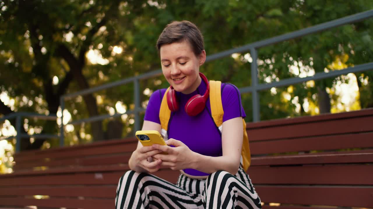 una chica con un corte de pelo corto en una camiseta púrpura, pantalones a rayas y una mochila amarilla se sienta en puestos marrones y habla en un teléfono amarillo