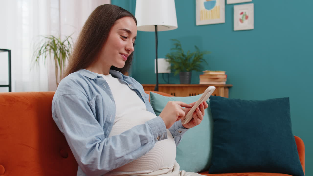 스마트폰을 사용하는 미소 짓는 임신한 여성이 가정 소파에 앉아있는 아기를 위해 온라인 구매를합니다.